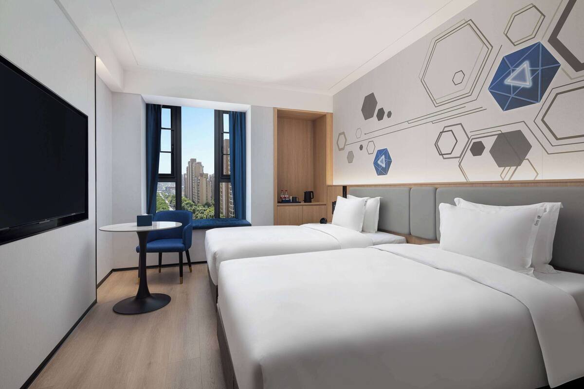 Een hotelkamer met twee bedden, een bureau, een stoel en een groot raam met uitzicht op gebouwen.