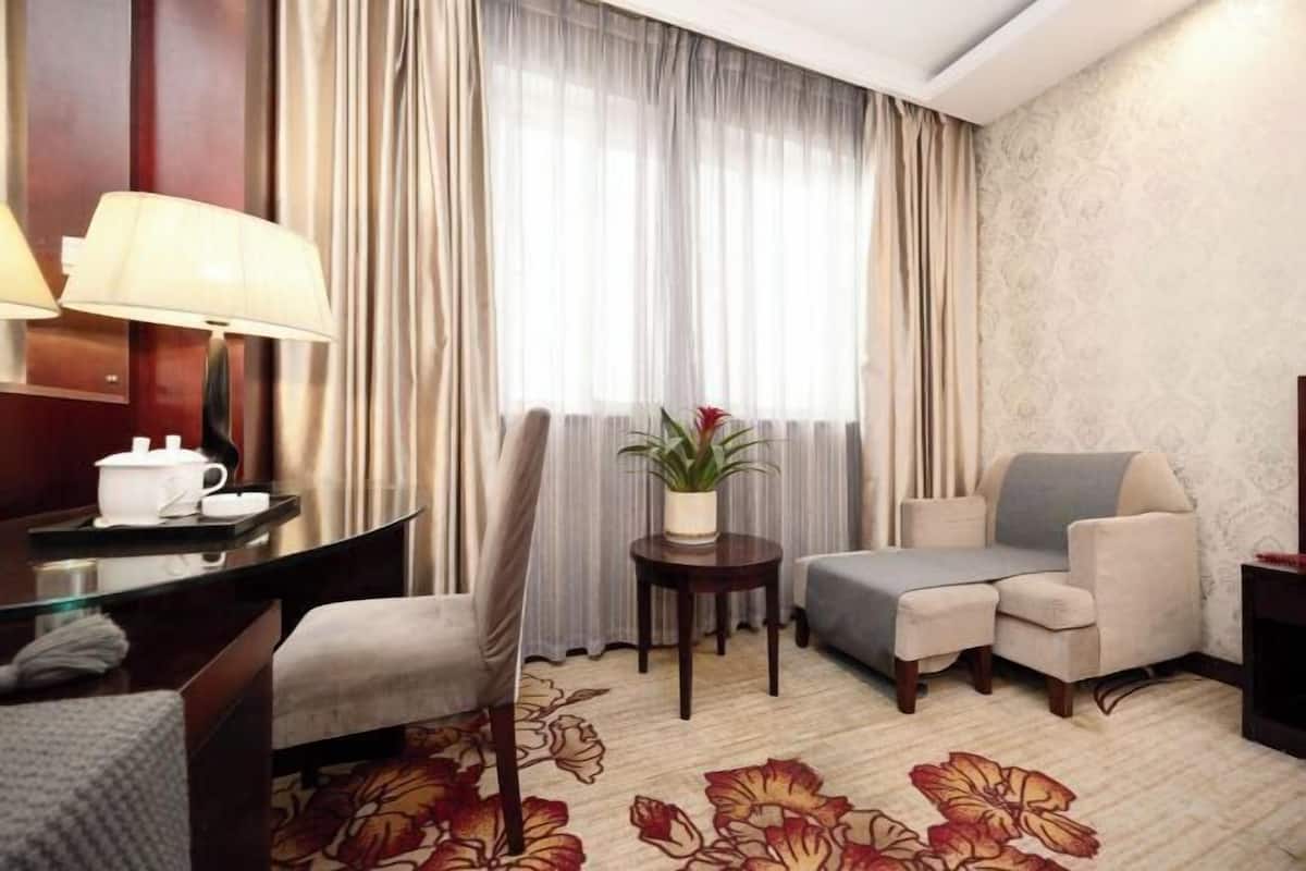 Habitación de hotel con escritorio, dos sillas, sofá, una mesita con planta y ventana con cortinas.