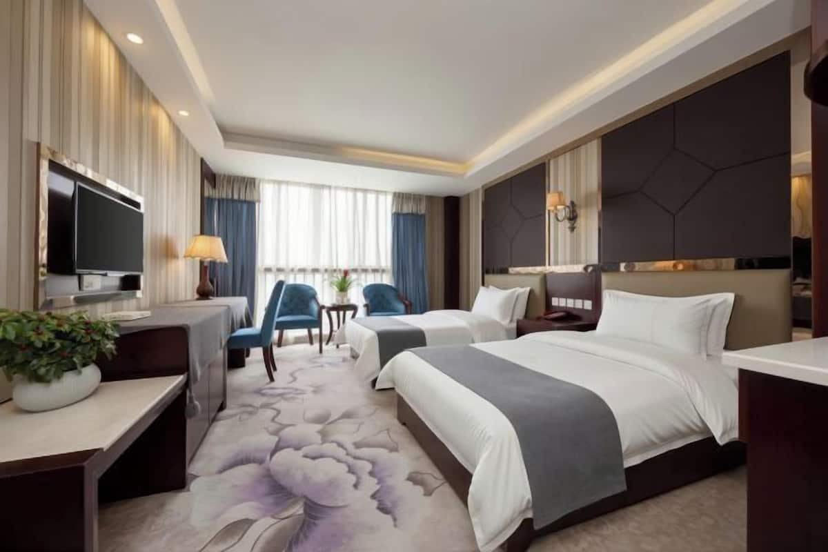 Habitación de hotel con dos camas, un escritorio con televisor, una pequeña mesa con sillas y un ventanal con cortinas.