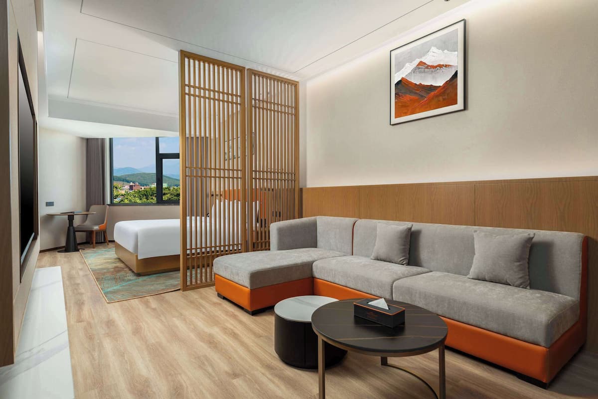 Een moderne hotelkamer met een hoekbank, een klein rond tafeltje, een houten vloer, uitzicht op bergen en een schuifdeur.