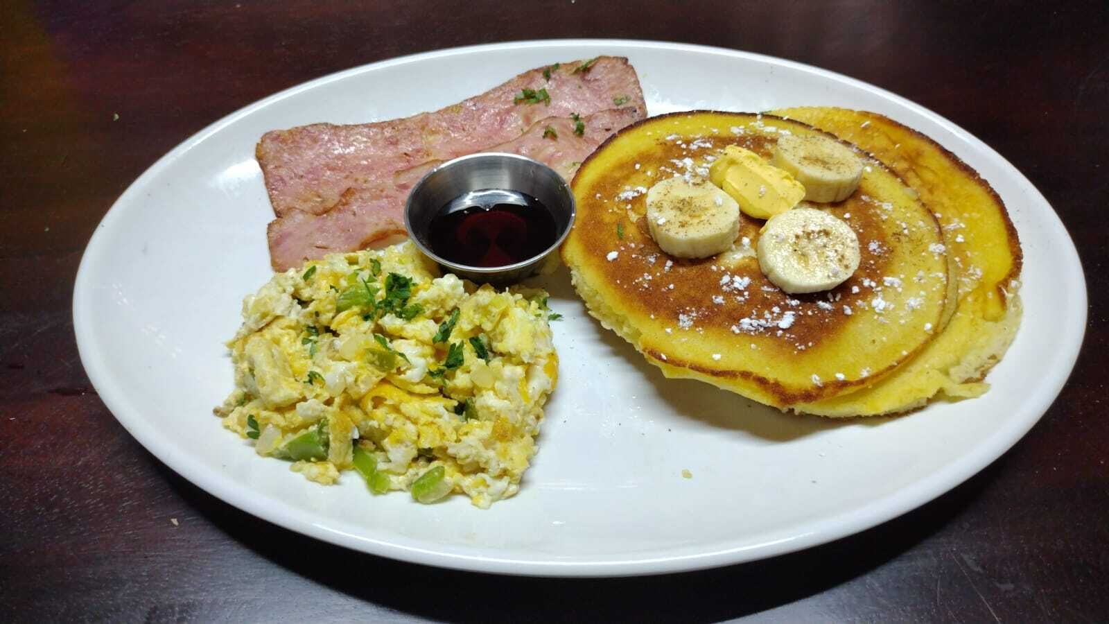 Un plato con huevos revueltos, tocino, panqueques cubiertos con rodajas de plátano y un recipiente pequeño de jarabe.