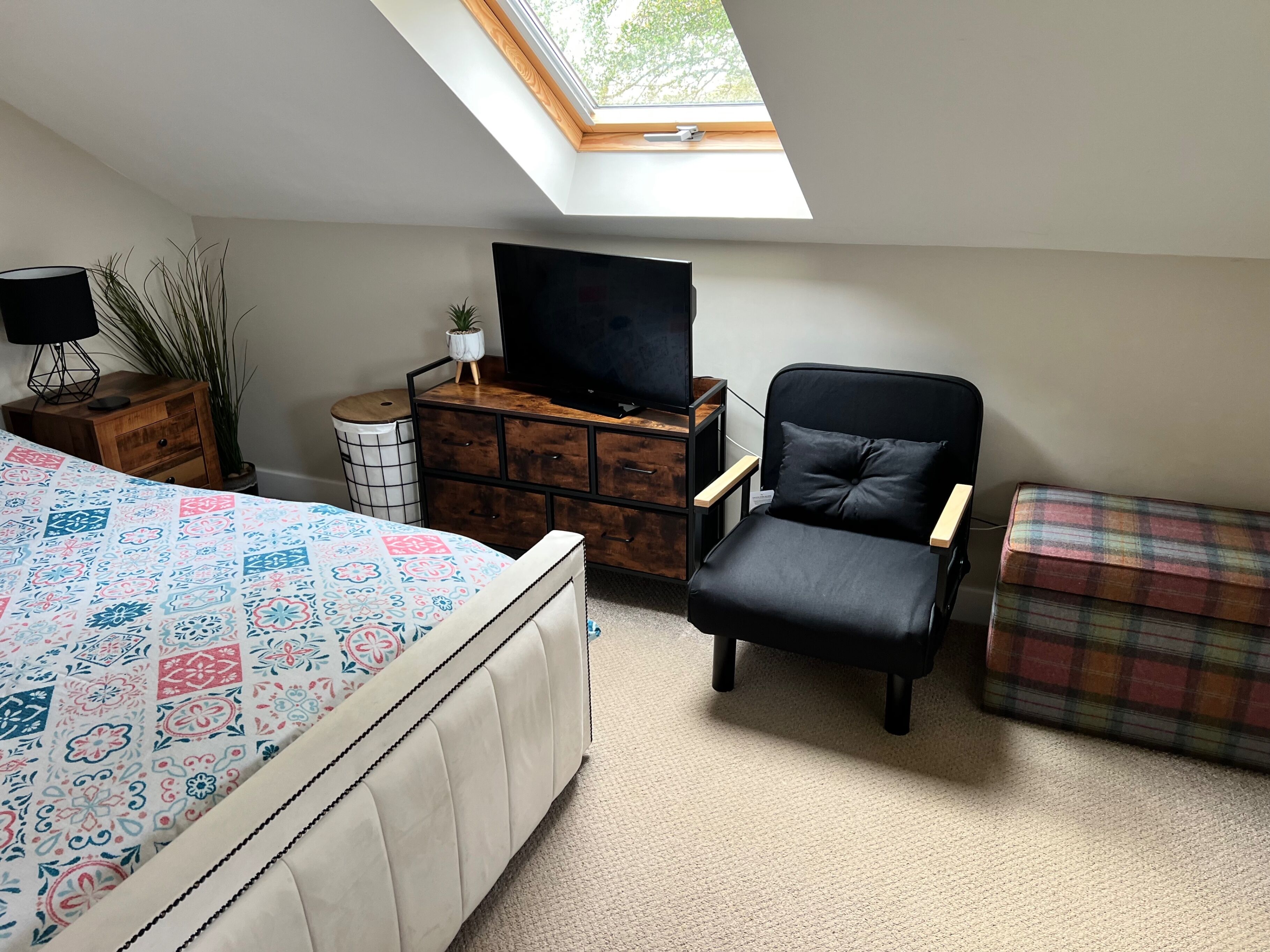 A bedroom with a bed, a TV, a chair, a small table, and a window.