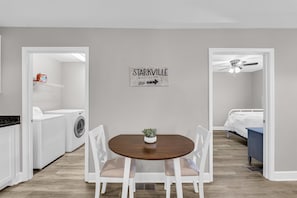 Una habitación con una mesa de comedor de madera, sillas blancas, una pequeña planta en maceta, un letrero en la pared, una lavadora, una cama y un ventilador de techo.
