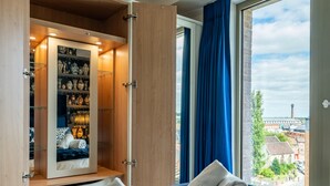 Un dormitorio con un ventanal grande, una cama con almohadas, un armario con estantes y vistas a edificios.