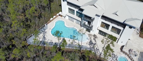 An aerial view of a modern house with a large swimming pool, surrounded by a forested area.