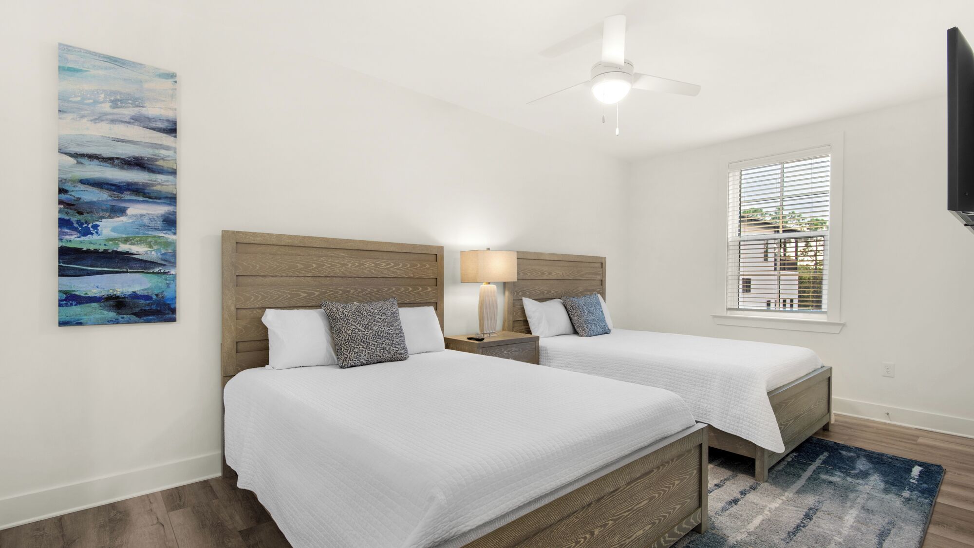 Habitación de hotel con dos camas, ventilador de techo, ventana con persianas y un cuadro en la pared.