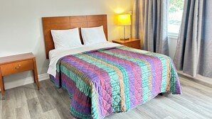 A neatly made bed with a colorful quilt, a wooden nightstand with a lamp, and a window with curtains. - Brant Inn (Brantford)
