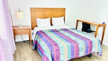 Una habitación de hotel con cabecera de madera, una cama con colcha colorida, una mesita de noche con lámpara y un escritorio con silla.