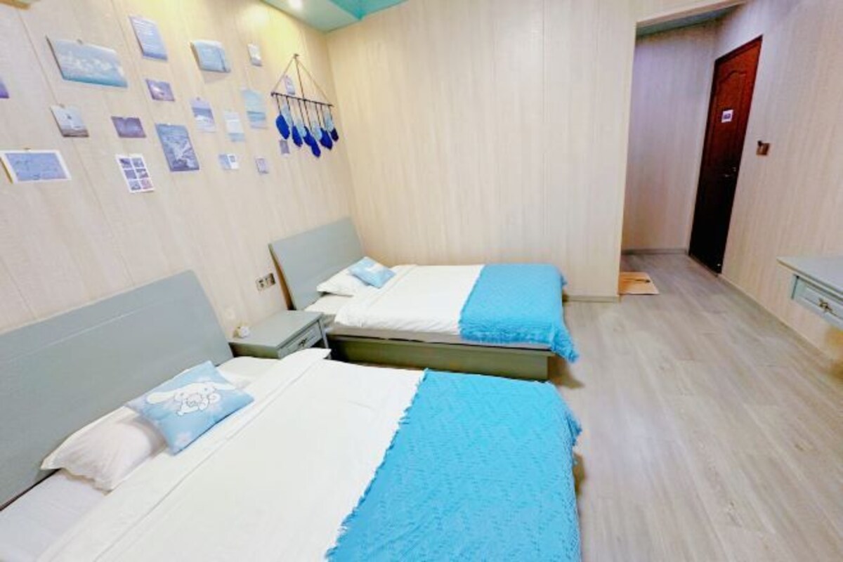 A room with two beds, a small bedside table, a wall-mounted shelf, and a door.