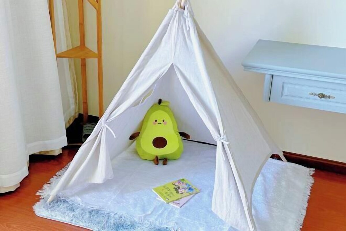 A tent-like structure with a plush toy inside, a small book on the floor, and a white shelf against the wall.