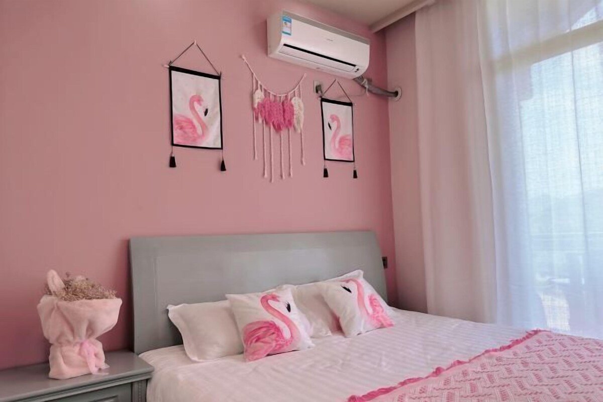 A bedroom with a pink wall, a bed with white bedding and pink flamingo pillows, a bedside table with a vase of dried flowers, and an air conditioning unit mounted on the wall.