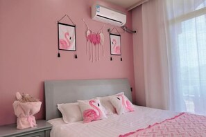 A bedroom with a pink wall, a bed with white bedding and pink flamingo pillows, a bedside table with a vase of dried flowers, and an air conditioning unit mounted on the wall. - Qiaoyu Inn (Zhoushan)