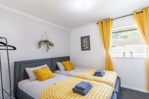 A hotel room with two beds, a hanging plant, a framed artwork, and a window with yellow curtains. - Open Mind Property - 4 Beds 2 Bathrooms & Parking (Portsmouth)