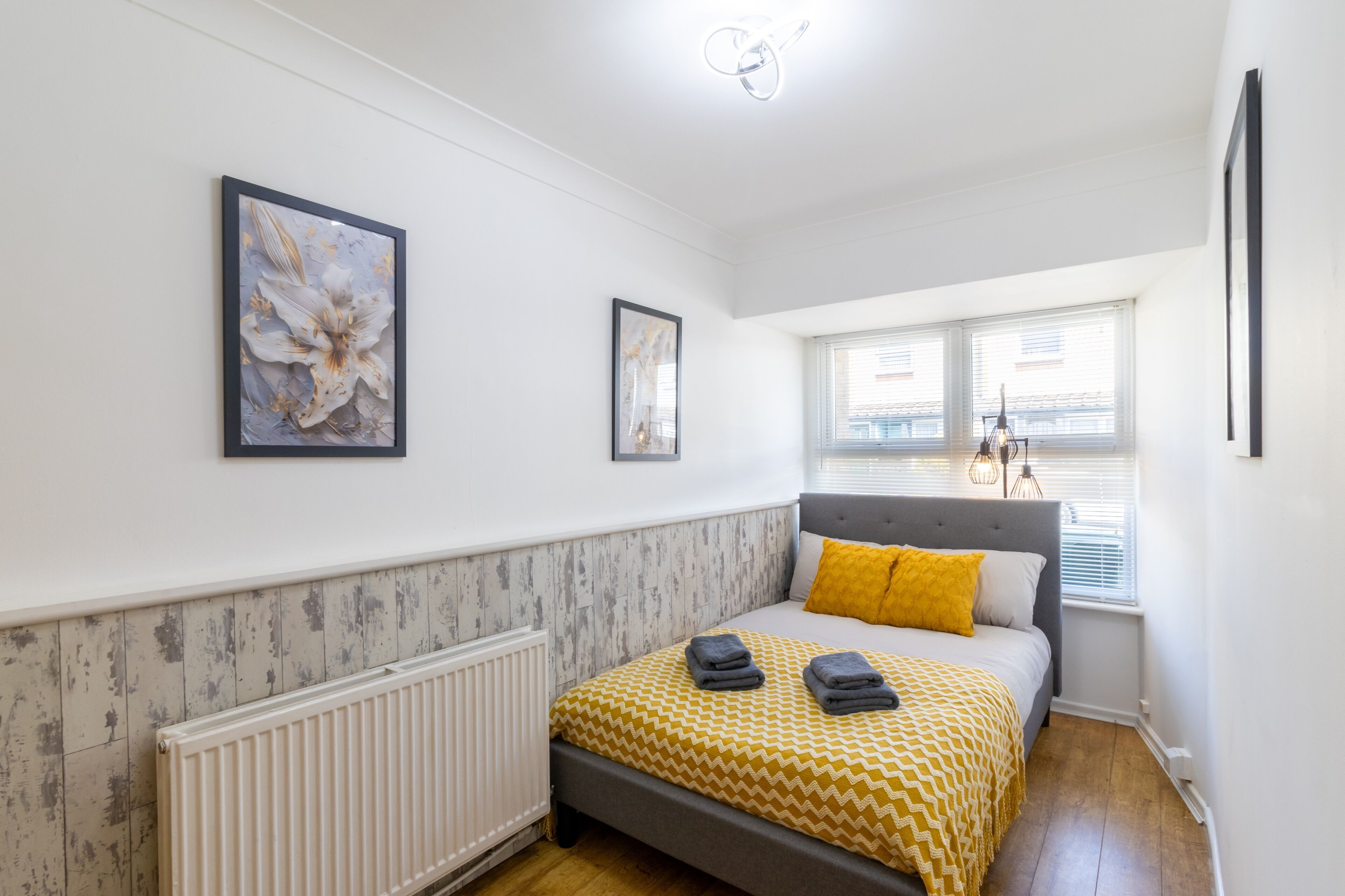 Ein Schlafzimmer mit einem Bett, zwei gerahmten Kunstwerken, einem Heizkörper und einem Fenster mit Jalousien.