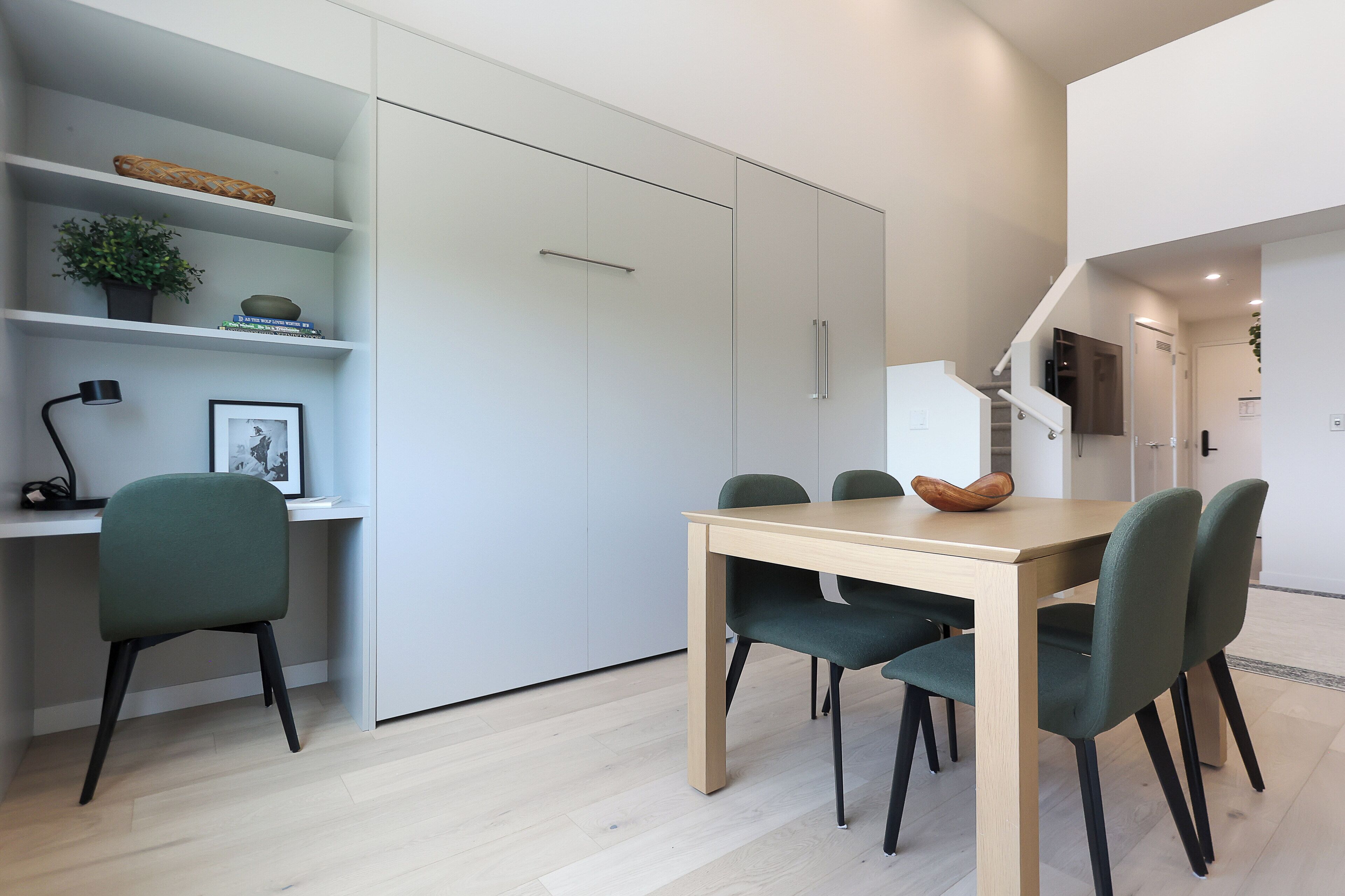 Een moderne eethoek met een houten tafel en vier stoelen, een boekenkast met decoratieve items, en een trap die naar een ander niveau leidt.