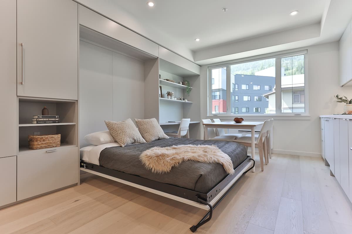 A modern bedroom with a bed, built-in storage, a desk, and a window with a view of buildings.
