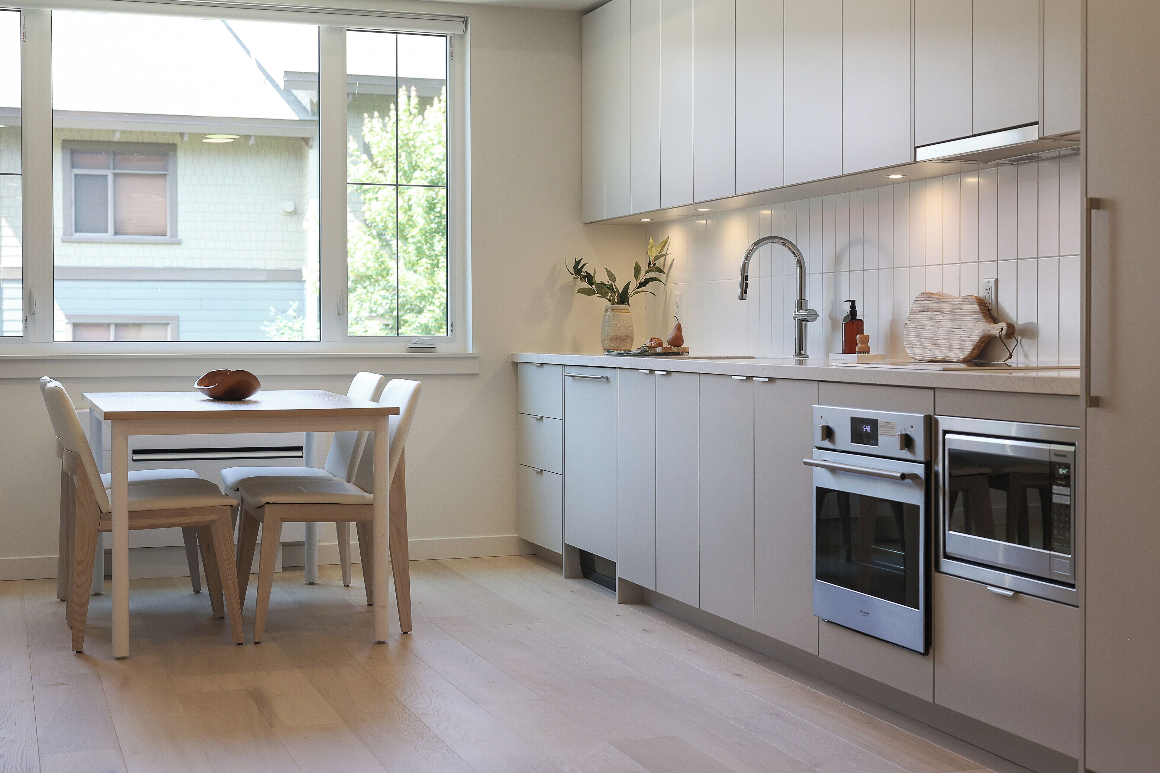 A modern kitchen with white cabinets, stainless steel appliances, a dining table with chairs, and a window with a view of a house.