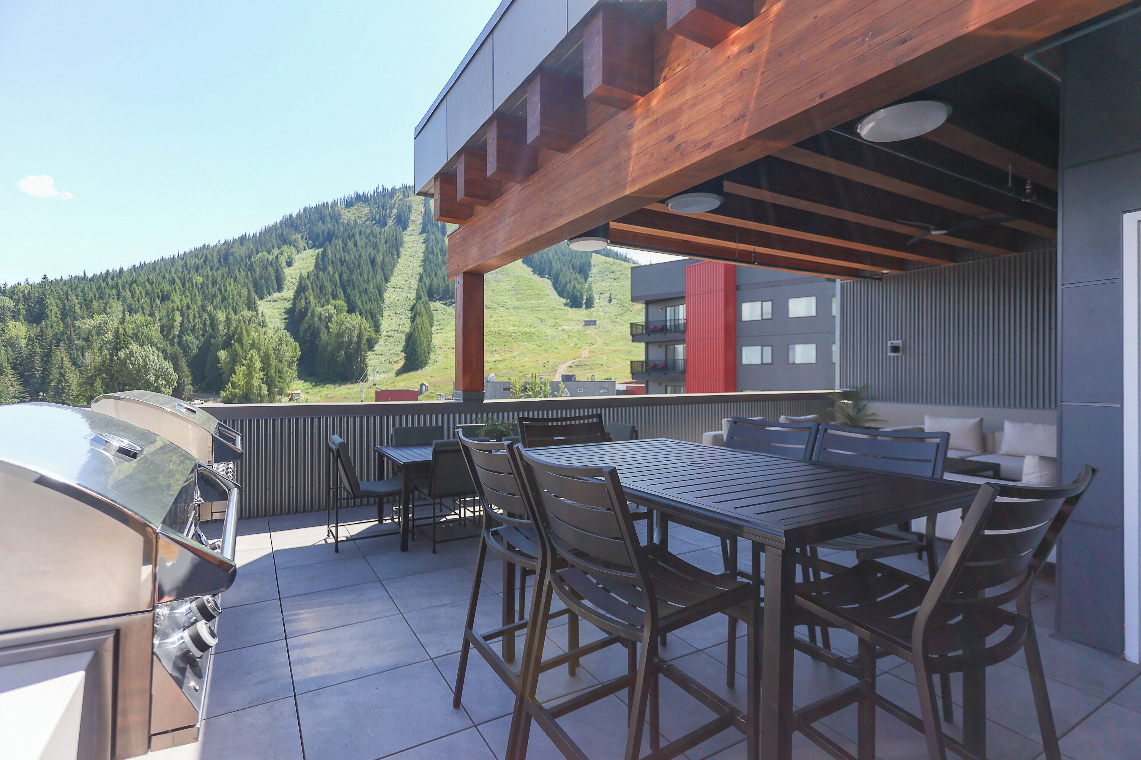 An outdoor patio with a wooden table and chairs, a barbecue grill, and a view of a green hillside.