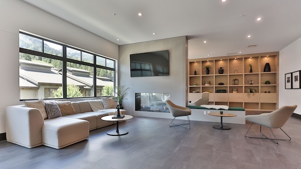 A modern living room with a sectional sofa, a round coffee table, and two chairs. There is a fireplace, a large window, and built-in shelving with decorative items. - The Crescent (Rossland)
