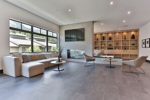 Ein modernes Wohnzimmer mit einem Ecksofa, einem runden Couchtisch und zwei Sesseln. Ein Kamin, ein großes Fenster und ein Einbauschranksystem mit dekorativen Gegenständen sind ebenfalls vorhanden.