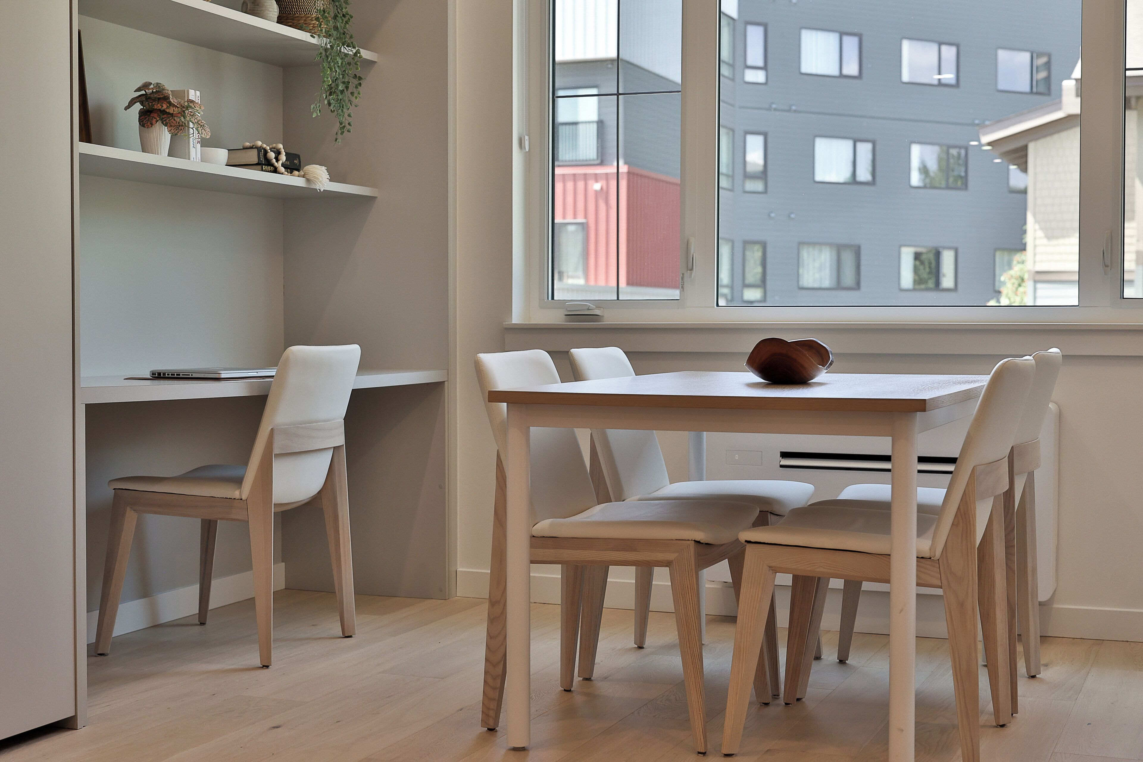 A modern dining area with a white table and chairs, a shelf with decorative items, and a window with a view of buildings.