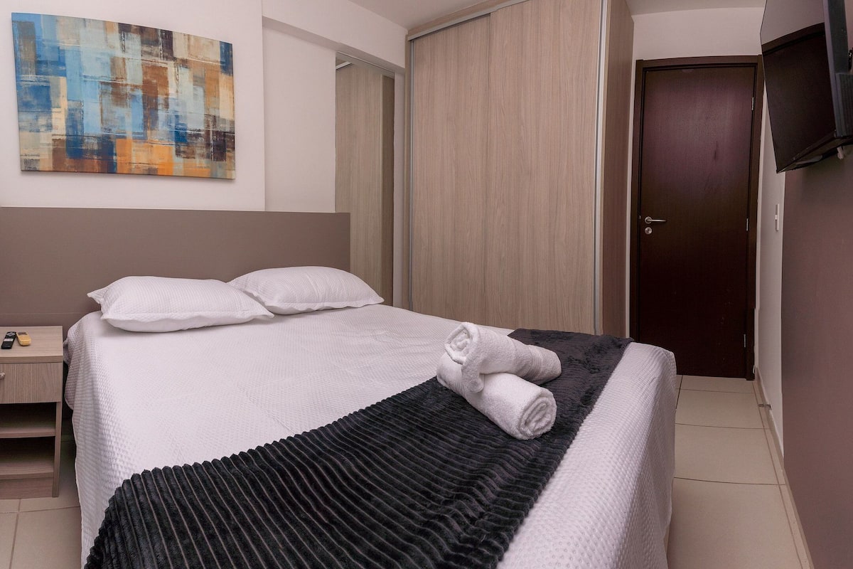 A neatly made bed with white bedding and a folded towel, a wooden nightstand with a phone, a painting on the wall, and a closed door.