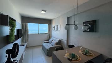 Una sala de estar moderna con un sofá gris, una mesa de comedor con una copa de vino y una taza, y un ventanal con vista al océano.