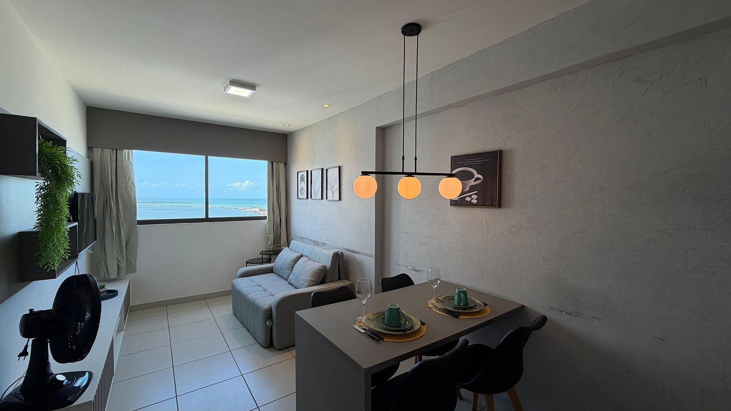 Ein modernes Wohnzimmer mit einer Couch, einem Esstisch für zwei Personen und einem großen Fenster mit Blick auf das Meer.