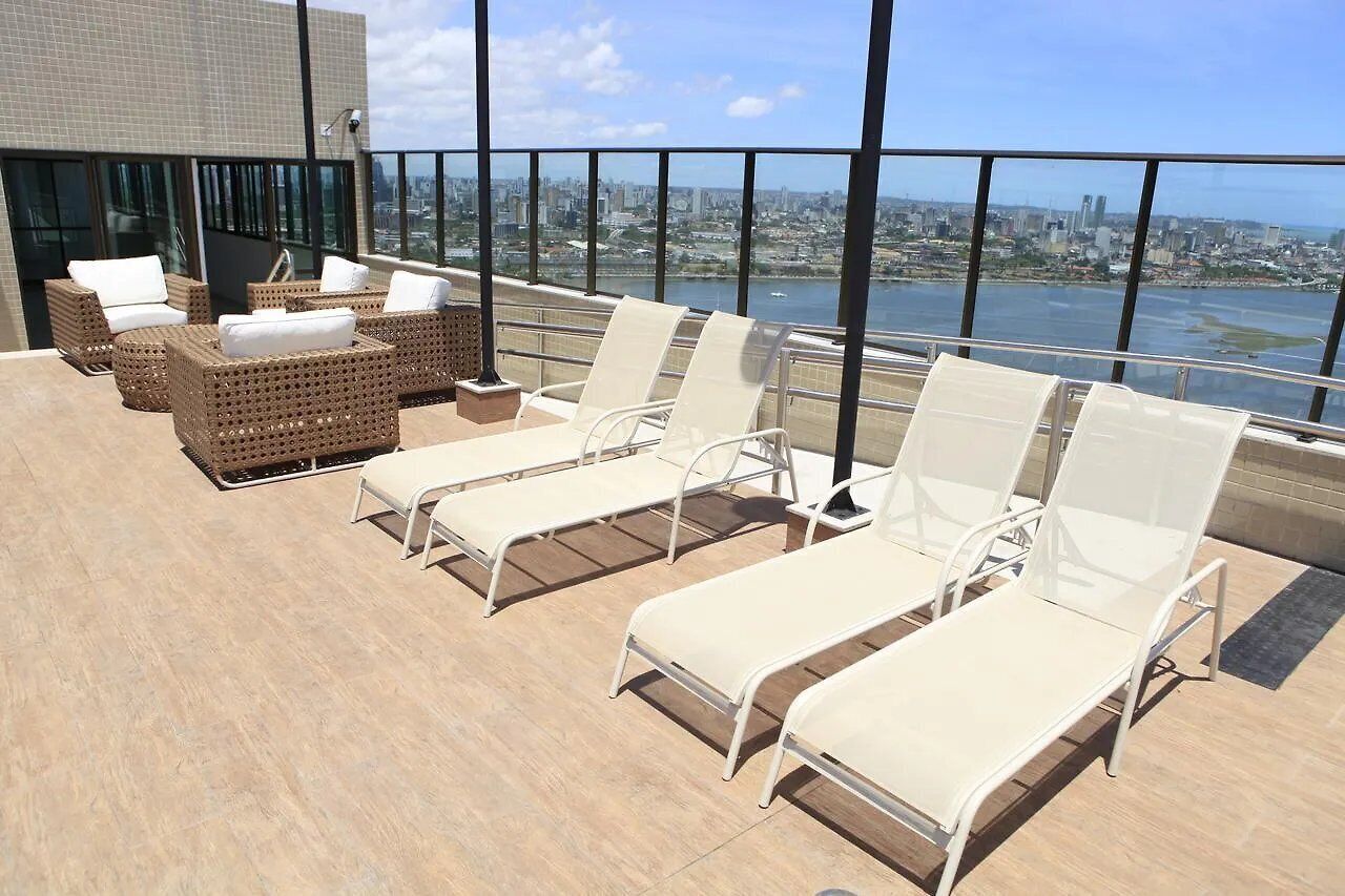 A rooftop lounge with wicker furniture and white lounge chairs, overlooking a cityscape and water.