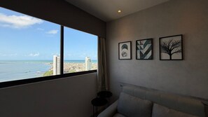 Una habitación con un ventanal grande que ofrece vistas a la ciudad y al mar, un sofá gris y tres obras de arte enmarcadas en la pared.