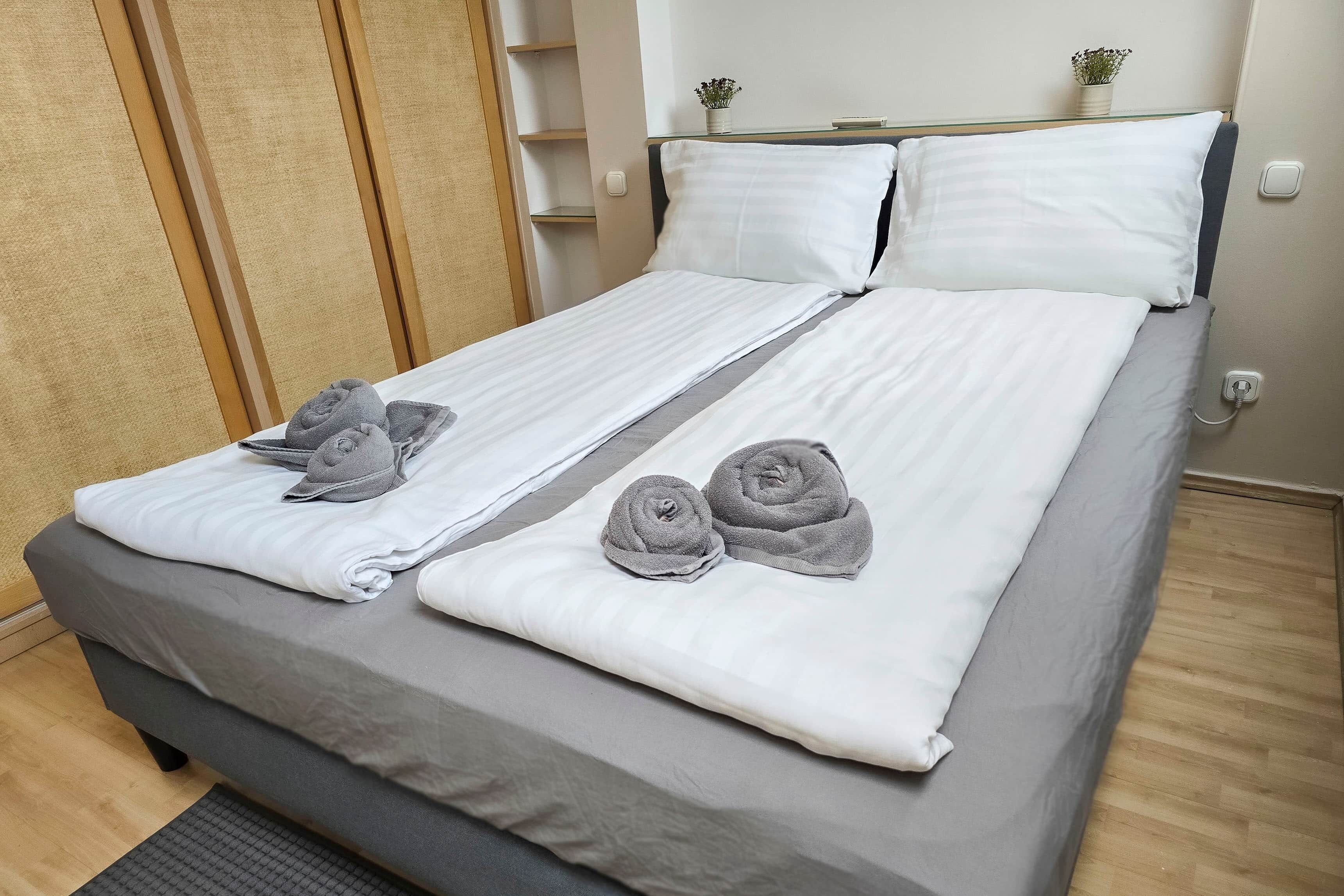 A double bed with grey and white bedding, two pillows, and two rolled towels.