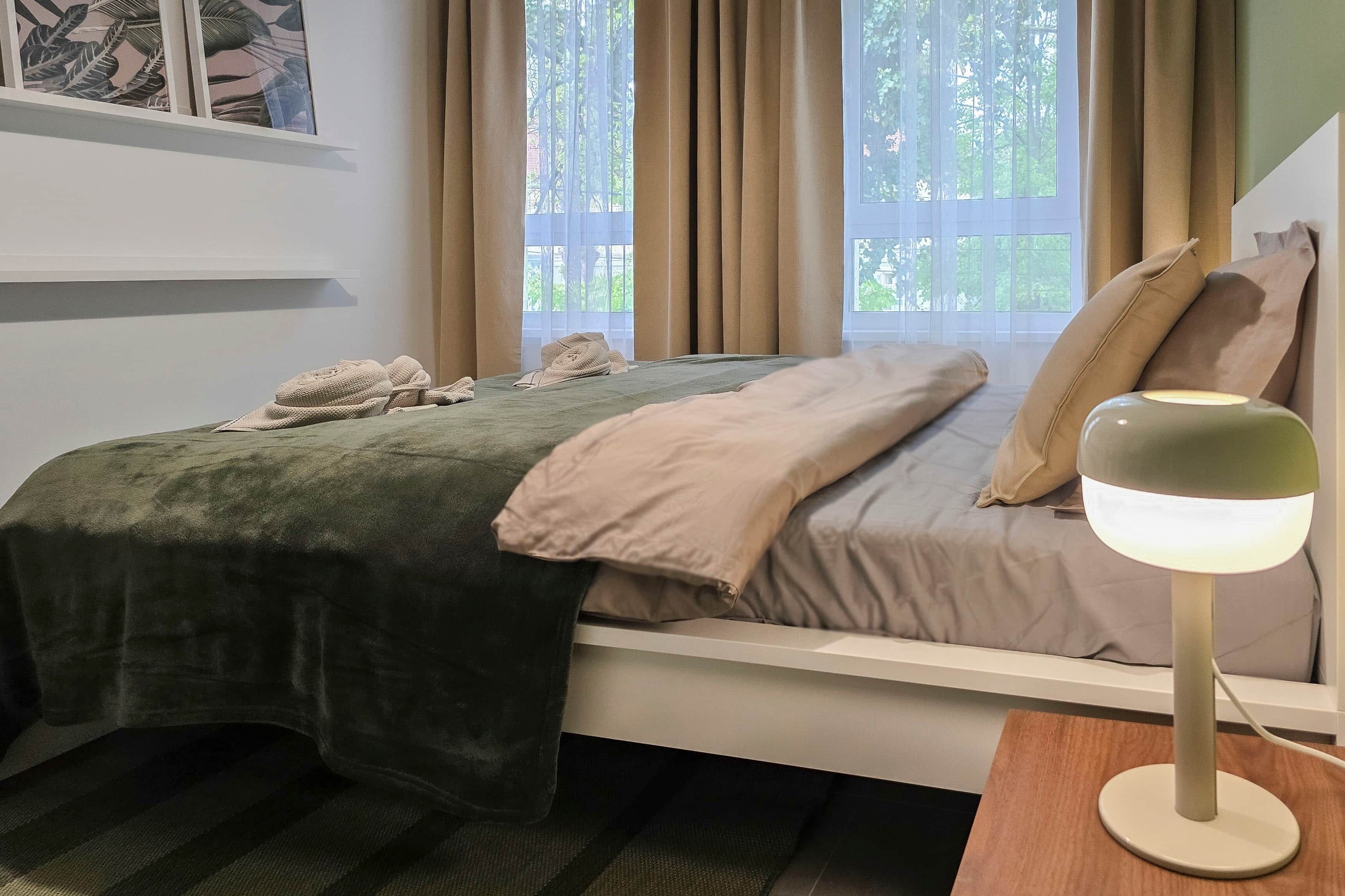 Et soveværelse med en seng, et natbord med en lampe, et vindue med gardiner og en væg med et billedramme.
