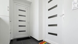 A hallway with a white door featuring multiple black horizontal slits, a tiled floor, and a colorful circular rug at the entrance. - IMCT - Green Home Apartment (Törökbálint)