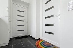 A hallway with a white door featuring multiple black horizontal slits, a tiled floor, and a colorful circular rug at the entrance. - IMCT - Green Home Apartment (Törökbálint)