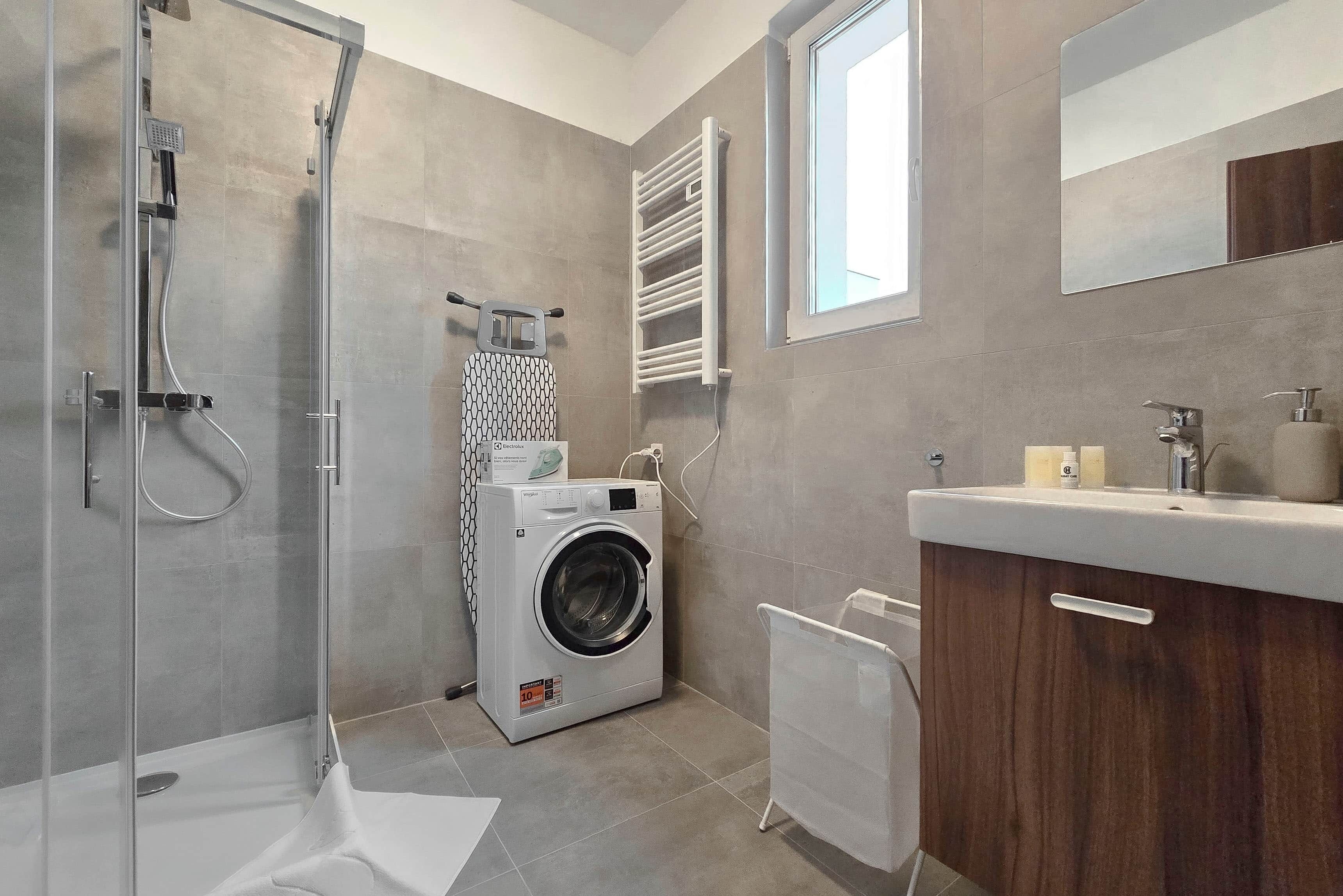A modern bathroom with a glass shower enclosure, a built-in washing machine, and a wooden vanity with a mirror.