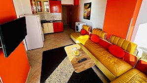 A modern living room with a sofa, a flat-screen TV, and a kitchen area with red cabinets and a washing machine. - Beachfront Apartment With Stunning Sea Views (Durrës)
