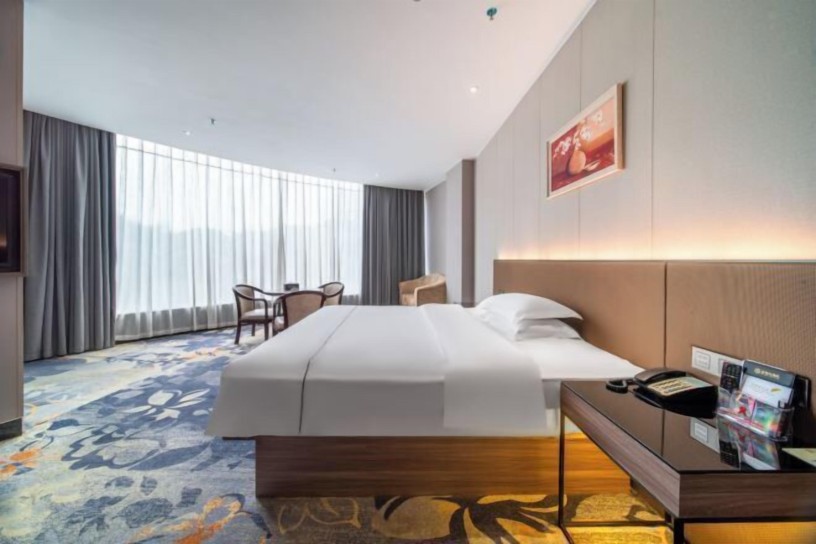 Een hotelkamer met een groot bed, een bureau met telefoon en tijdschriftenrek, een stoel en een groot raam met gordijnen.
