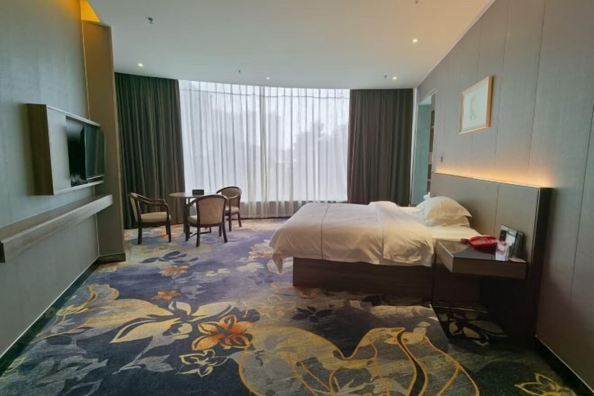 A hotel room with a bed, a desk, a chair, a TV, and a large window with curtains.