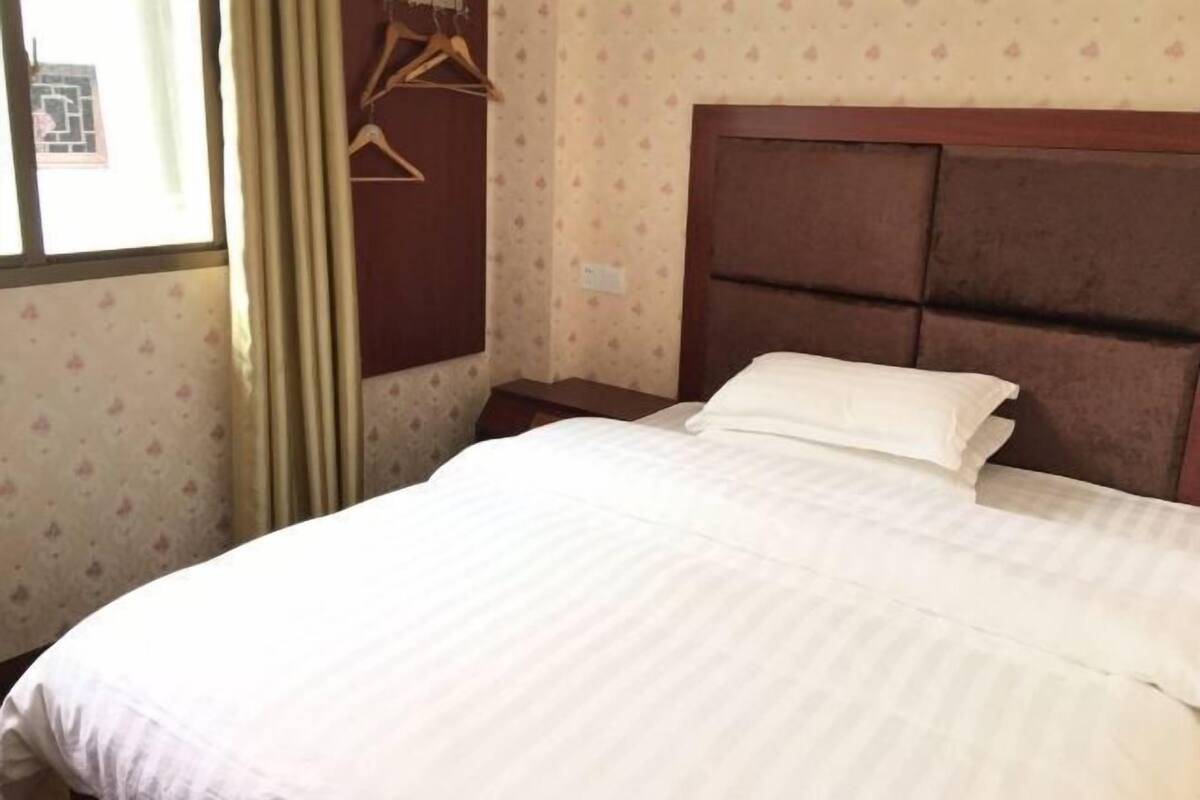 Una cama bien hecha con cabecera, una mesita de noche de madera y un ventanal con cortinas.