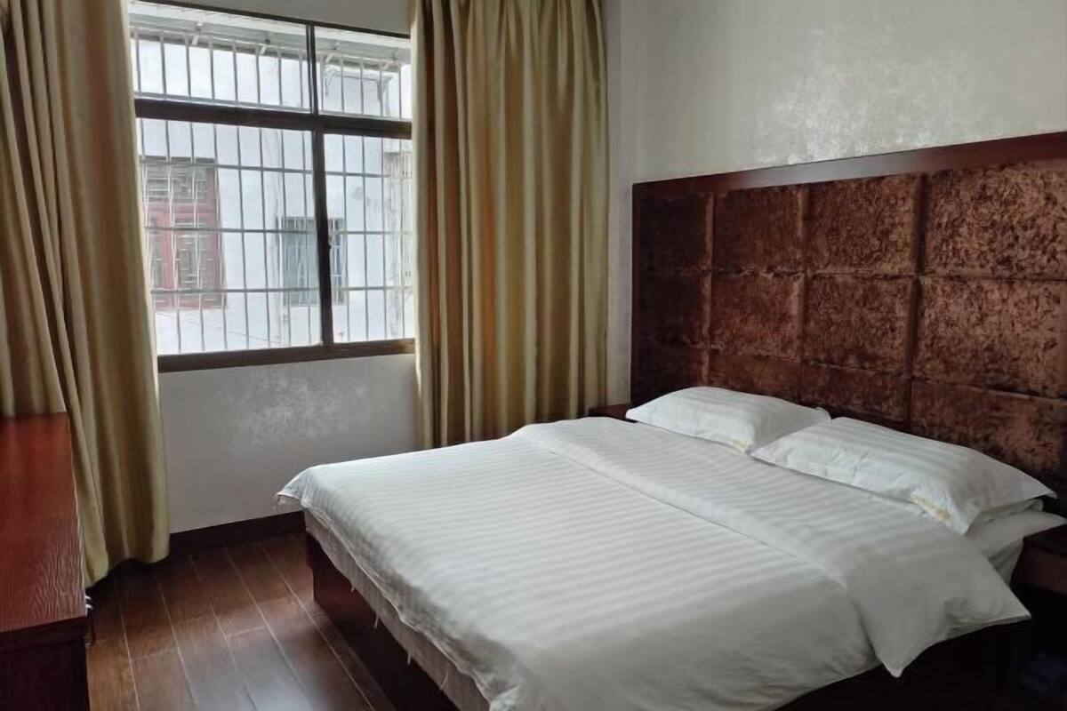 Habitación de hotel con una cama grande, cabecera de madera, cortinas y una ventana con barrotes.