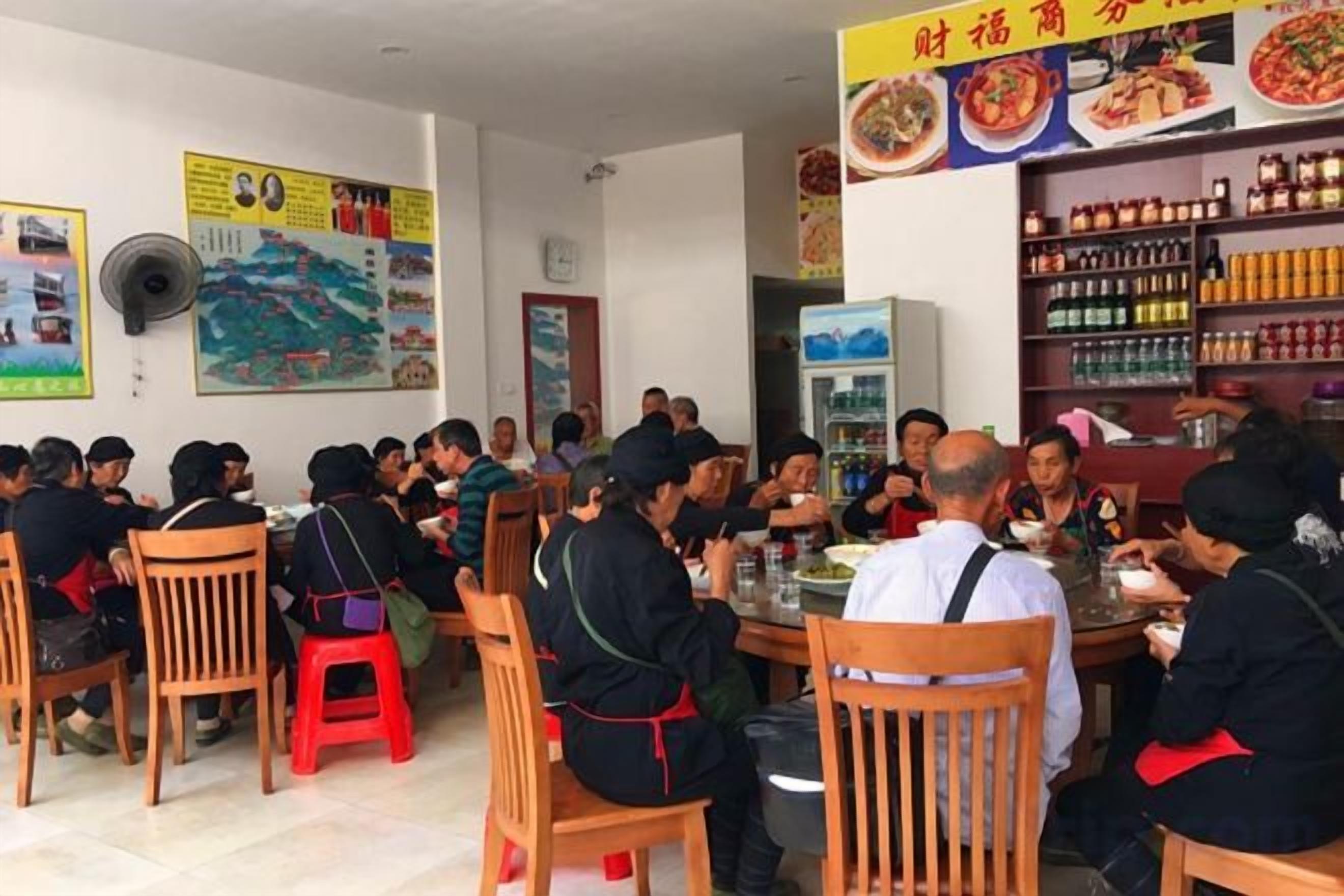 A group of people dining in a restaurant with wooden chairs and tables, a wall with posters, and shelves with bottles.
