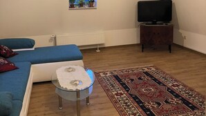 Una camera mansardata accogliente con soffitto in legno, un televisore su un mobile, un divano blu, un tavolino con tovaglia in pizzo e un tappeto decorato.