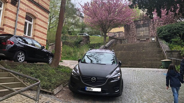 Un'auto nera parcheggiata su un vialetto lastricato in una zona residenziale con edifici in mattoni, una scala e alberi in fiore.