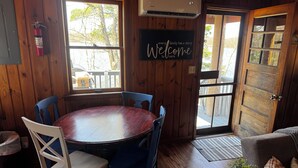 A wooden interior with a round table, blue chairs, a welcome sign, and a door leading outside.