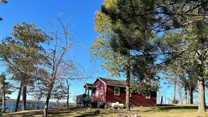 A red cabin with a porch, surrounded by trees and a lake.