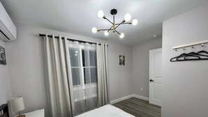 A bedroom with a bed, a window with curtains, a wall-mounted air conditioner, a ceiling light fixture, and a door. - SI HOUSE - Lovely 2 bedroom unit in Staten Island (Staten Island)