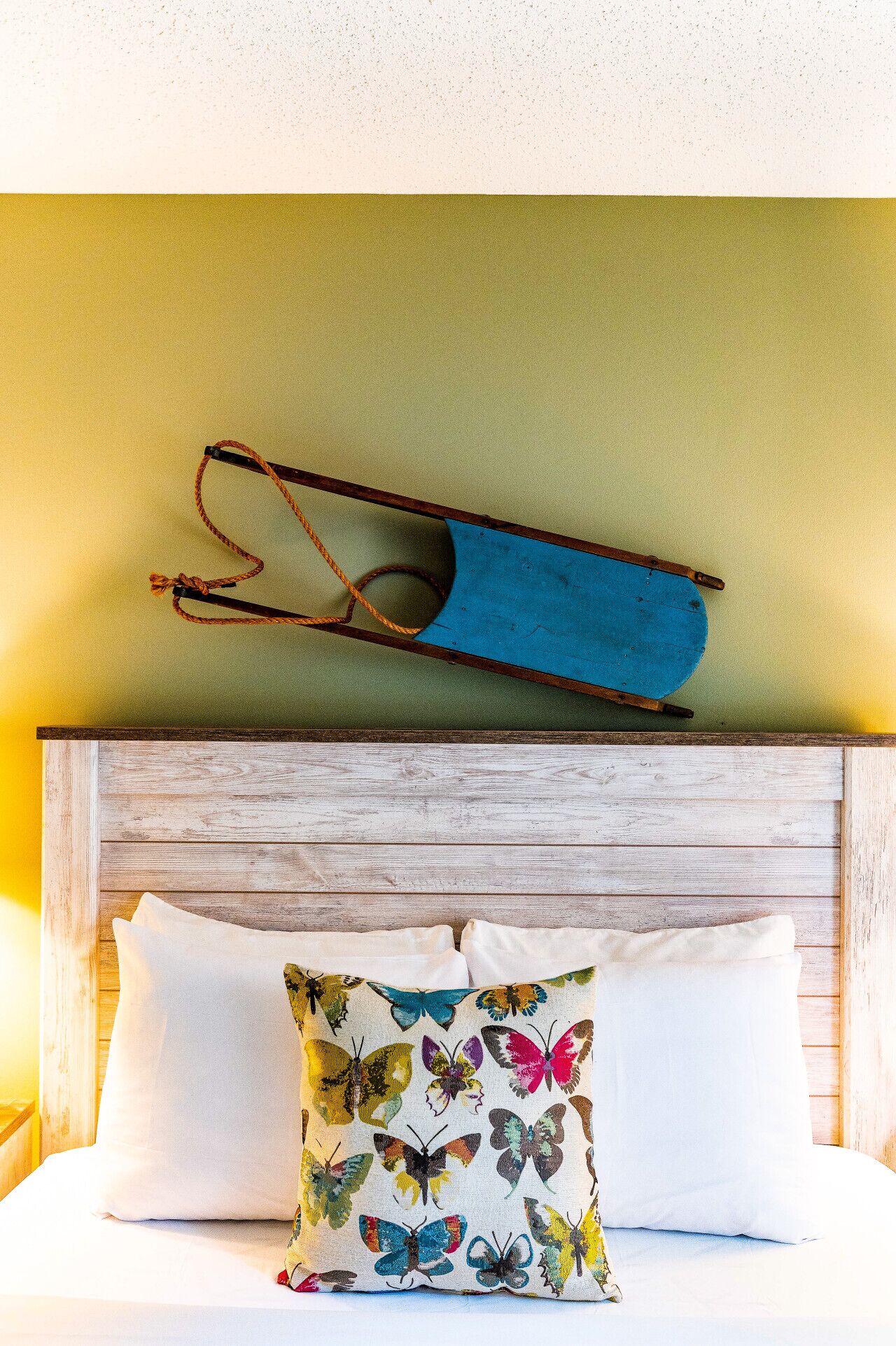 Un lit avec des oreillers blancs et un coussin coloré en forme de papillon, une tête de lit en bois et un objet bleu suspendu au-dessus du lit.
