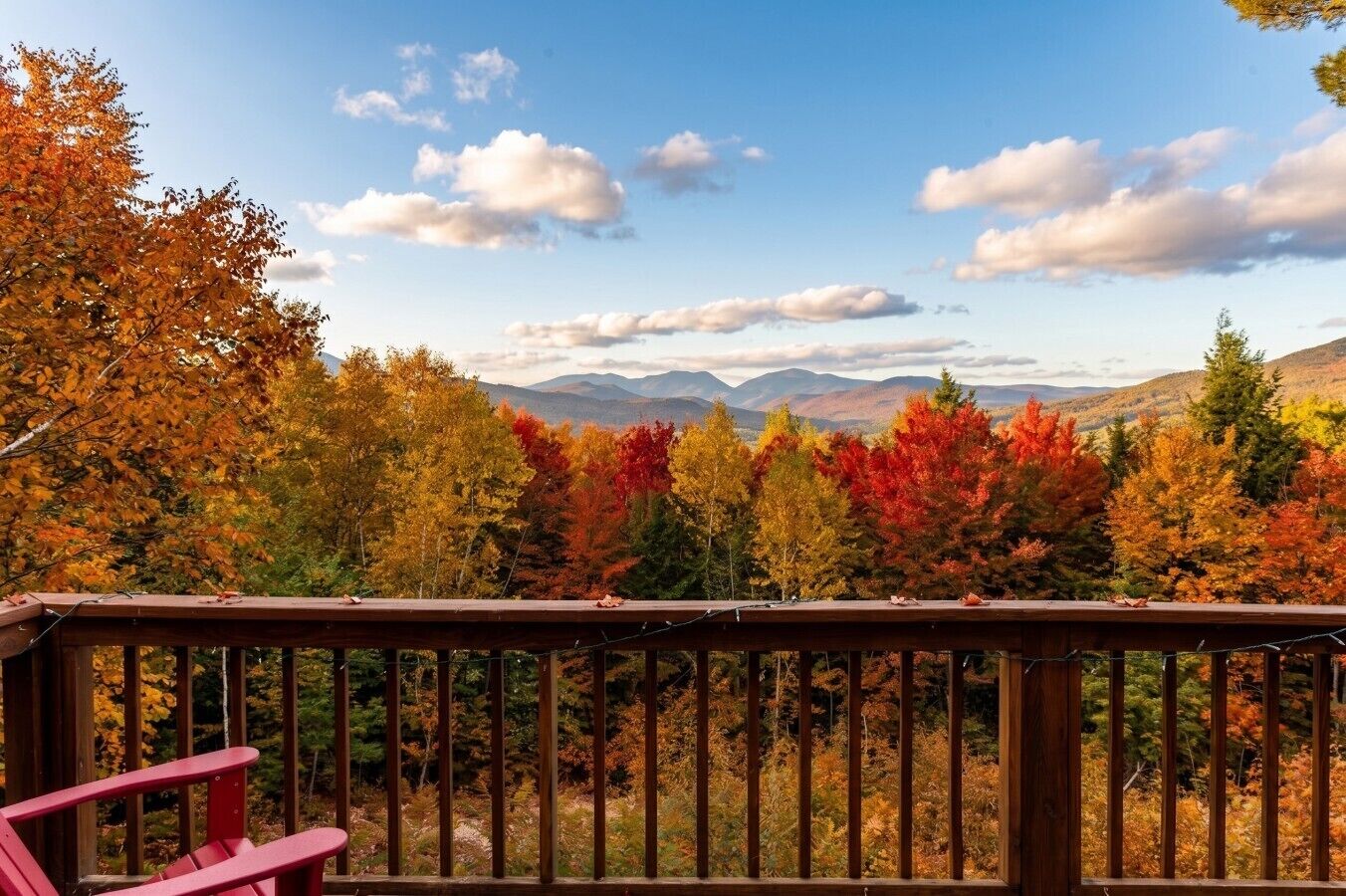 Una terraza de madera con una silla Adirondack roja, con vistas a un bosque de follaje otoñal y montañas distantes.