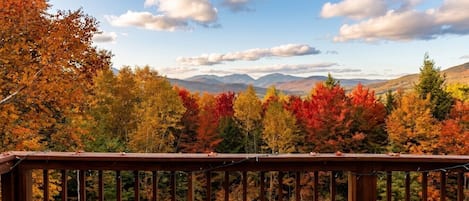 Una terraza de madera con una silla Adirondack roja, con vistas a un bosque de follaje otoñal y montañas distantes.