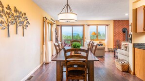 A dining area with a wooden table and chairs, a chandelier, and a view of the outdoors through large windows. - Attitash Awesomeness - Fun for the Whole Family (Bartlett)